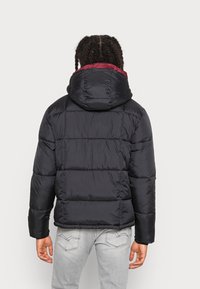 Černá péřová bunda s kapucí, s bordó podšívkou, prošívaným vzorem a elastickými manžetami, nošená přes světle šedé džíny.