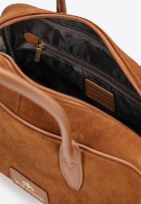 Borsa in suede marrone con manici in pelle, chiusura con zip e foderatura interna scura. Presenta una tasca con zip e un'etichetta in pelle brandizzata all'interno.