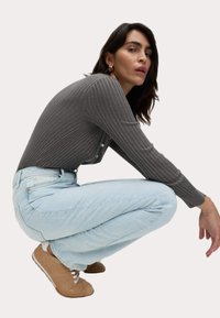 Cardigan grigio a costine con bottoni, abbinato a jeans azzurri chiari e sneakers beige. Il modello è accovacciato, mostrando la vestibilità e le trame dell'outfit.