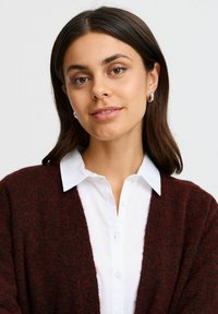 Cardigan lavorato a maglia color bordeaux sopra una camicia bianca con colletto, caratterizzato da una vestibilità comoda e un tessuto testurizzato. Il modello posa con un'espressione neutra.