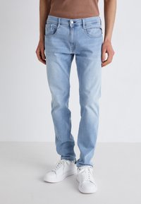 Vaqueros de denim azul claro con corte slim, que cuentan con bolsillos frontales, un cierre de cremallera y botón, y una sutil textura desgastada. Llevados con zapatillas blancas.