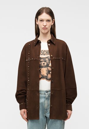 Vrouw draagt een oversized bruine jas met studs over een wit t-shirt met een teddybeerprint en lichtblauwe spijkerbroek, staand tegen een effen achtergrond.