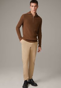 Brauner gerippter Strickpullover mit Reißverschlusskragen, kombiniert mit hellbeigen Hosen und schwarzen Schnürschuhen. Einfaches Design mit strukturiertem Stoff.