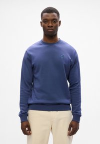 Temno modra majica s kapuco in okroglim izrezom, z rebrastimi manšetami in spodnjim robom. Ima majhen, kvadratni logotip na prsih. Gladka tekstura.