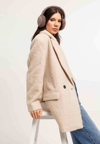 Cappotto beige a trama con ampio colletto, due tasche frontali e due bottoni neri. Il modello indossa cuffie morbide grigie e si trova seduto su uno sgabello.