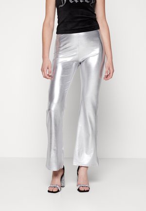 Kobieta ubrana w błyszczące, srebrne rozszerzane spodnie, czarną bluzkę i czarne sandały na wysokim obcasie z paskami zdobionymi kryształkami na białym tle.