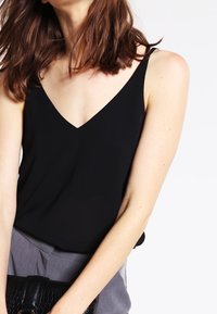 Svart V-ringad linne med tunna remmar. Slätt tyg, figursydd silhuett. Kombinerad med grå högmidjade byxor och en strukturerad svart handväska.