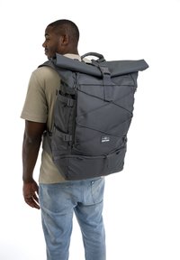 Mochila negra de tipo roll-top con correas ajustables, hecha de un tejido duradero. Presenta detalles con cordones elásticos y un parche con el logo en la parte frontal.
