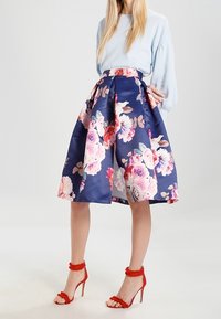 Kvinna i en ljusblå tröja, marinblå knä-lång kjol med stort rosa blommönster, och röda högklackade sandaler med ankelremmar.