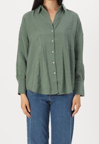 Chemise verte à boutons avec un tissu doux et texturé, dotée d'un col pointu et de manches longues, assortie à un jean bleu.