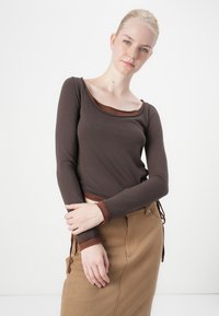 Brauner langärmliger Pullover mit rundem Ausschnitt, der einen kontrastierenden dunkleren braunen Saum am Hals und an den Bündchen hat, kombiniert mit einem beigen Rock.