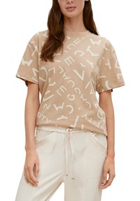 Beige bomulds t-shirt med geometrisk hvid bogstavtryk, korte ærmer, rund halsudskæring og en afslappet pasform. Parret med lyse bukser.