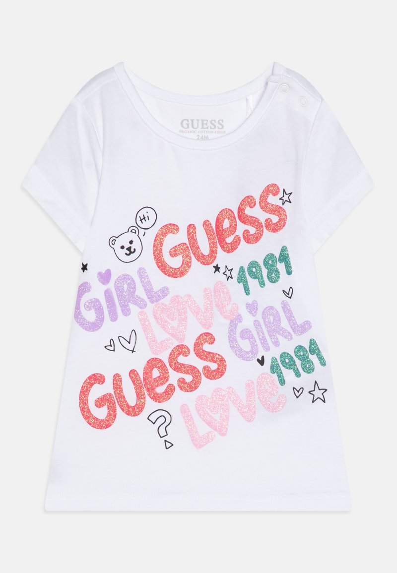 T-shirt blanc pour tout-petit avec texte pailleté coloré indiquant "Guess", "Girl", "Love", "1981", et petits cœurs, étoiles ainsi qu’un ours avec une bulle "Hi".