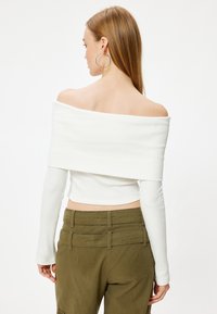 Weiße gerippte Off-Shoulder-Crop-Top mit langen Ärmeln, kombiniert mit olivgrünen hoch taillierten Hosen mit mehreren Gesäßtaschen und Nähdetails.