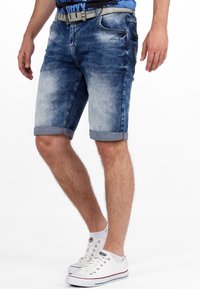 Denimshorts met een vervaagde blauwe wassing, opgerolde boorden en subtiele versleten details. Gecombineerd met witte sneakers en een grijze riem.