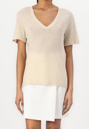 Personne portant un t-shirt beige à manches courtes avec col en V et une jupe blanche fendue à l'avant, debout devant un fond uni.