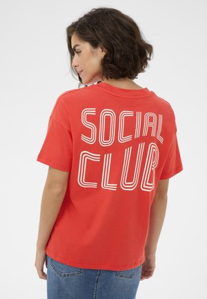 Jeune femme aux cheveux courts et bouclés portant un t-shirt rouge avec le texte blanc « SOCIAL CLUB » dans le dos, associé à un jean bleu.