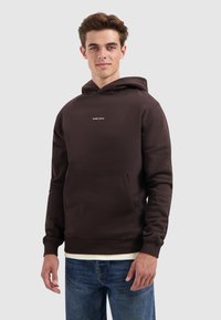 Jonge man in een donkerbruine hoodie met de tekst "PURE PATH" en blauwe jeans, staande tegen een effen lichte achtergrond.