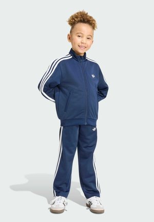 Child with curly hair stands smiling, wearing navy blue Adidas track jacket and pants with white stripes and white sneakers.