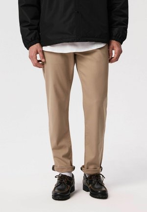 Mężczyzna w beżowych spodniach podwiniętych u dołu, czarnych skórzanych sznurowanych butach, białej koszuli i czarnej kurtce stojący na jednolitym tle.