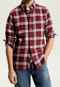 Homme portant une chemise à manches longues à carreaux rouges et blancs avec les manches retroussées, déboutonnée au col, associée à un jean bleu.