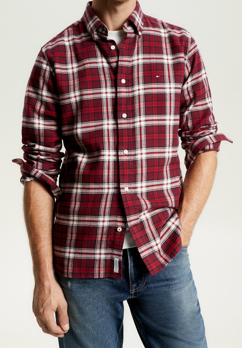 Homme portant une chemise à manches longues à carreaux rouges et blancs avec les manches retroussées, déboutonnée au col, associée à un jean bleu.