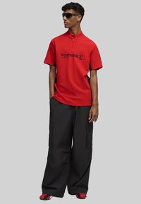 Camisa polo roja de manga corta con cuello de cremallera y paneles laterales negros, combinada con pantalones cargo negros de corte holgado y zapatos acentuados en rojo.