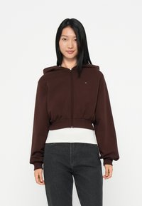 Brown cropped hoodie with a front zipper, wide sleeves, and a drawstring hood. Features a small logo on the chest and a soft texture.