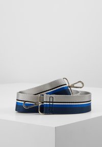 Cinturino per borsa in tessuto a righe grigio, nero, bianco, blu e navy con ganci a moschettone in metallo argentato su entrambe le estremità, arrotolato su una superficie bianca.