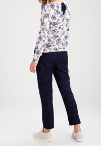 Frau trägt ein weißes, blumiges Langarmshirt mit blauen Blumen, marineblauen Nadelstreifenhosen und weißen Sneakers, stehend mit dem Rücken zur Kamera.