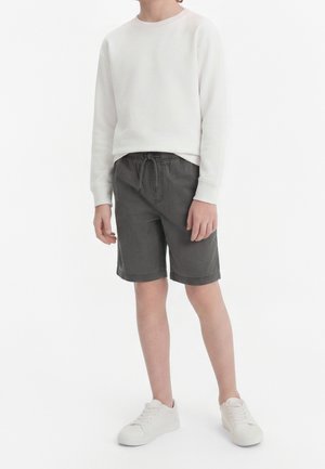 Enfant portant un sweat-shirt blanc à manches longues, un short gris foncé à cordon longueur genoux, et des baskets blanches, debout devant un fond blanc.