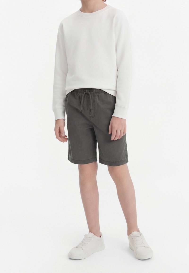 Enfant portant un sweat-shirt blanc à manches longues, un short gris foncé à cordon longueur genoux, et des baskets blanches, debout devant un fond blanc.