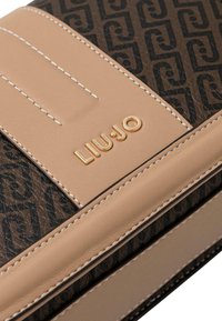 Cartera marrón con diseño estampado, detalles en cuero beige liso, costuras blancas y un logotipo dorado. Forma rectangular compacta con detalles texturizados.