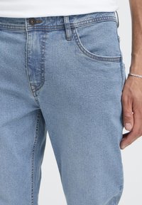 Svetlo modre kavbojke iz jeansa s klasičnim pet žepnim dizajnom, kovinskim gumbom za zapiranje in subtilno izbledenim vzorcem vzdolž stegnih in šivov.