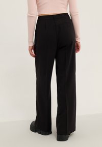 Pantalon noir taille haute, sur mesure, à jambes larges, avec une texture lisse, des plis subtils et des poches arrière. Complété par des chaussures noires épaisses.
