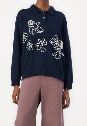 Maglione blu navy a maniche lunghe con colletto e bottoni, decorato con ricami floreali astratti bianchi, indossato con pantaloni larghi color malva.