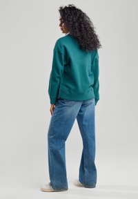 Felpa verde con una vestibilità rilassata, polsini e orlo a costine; abbinata a jeans in denim azzurro a vita alta e sneakers bianche.