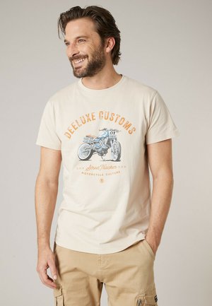 Homme souriant avec une barbe portant un t-shirt beige avec un motif de moto vintage et le texte "Deeluxe Customs Street Tracker Motorcycle Culture".