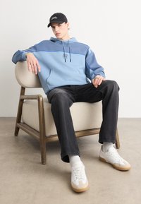 Zils hoody ar kontrastējošu tumši zilu paneli, ar šnurēm; melnas džinsas; baltas kedas ar beigu akcentiem; sēžot uz gaišas krēsla.