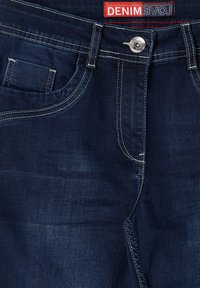 Vaqueros de mezclilla azul oscuro con un diseño clásico de cinco bolsillos, que cuentan con costuras blancas en contraste y un cierre de botón metálico en la cintura.