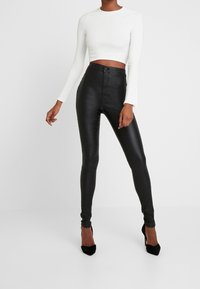 Svarta fusk läderleggings med hög midja, slät yta och knäppning framtill. Parad med en åtsittande vit långärmad topp.