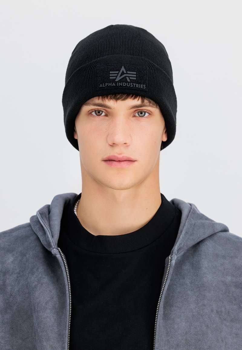 Młoda osoba z krótkimi brązowymi włosami nosi czarną czapkę Alpha Industries i szarą bluzę zapinaną na zamek na czarnej koszulce, patrząc na wprost z neutralnym wyrazem twarzy.