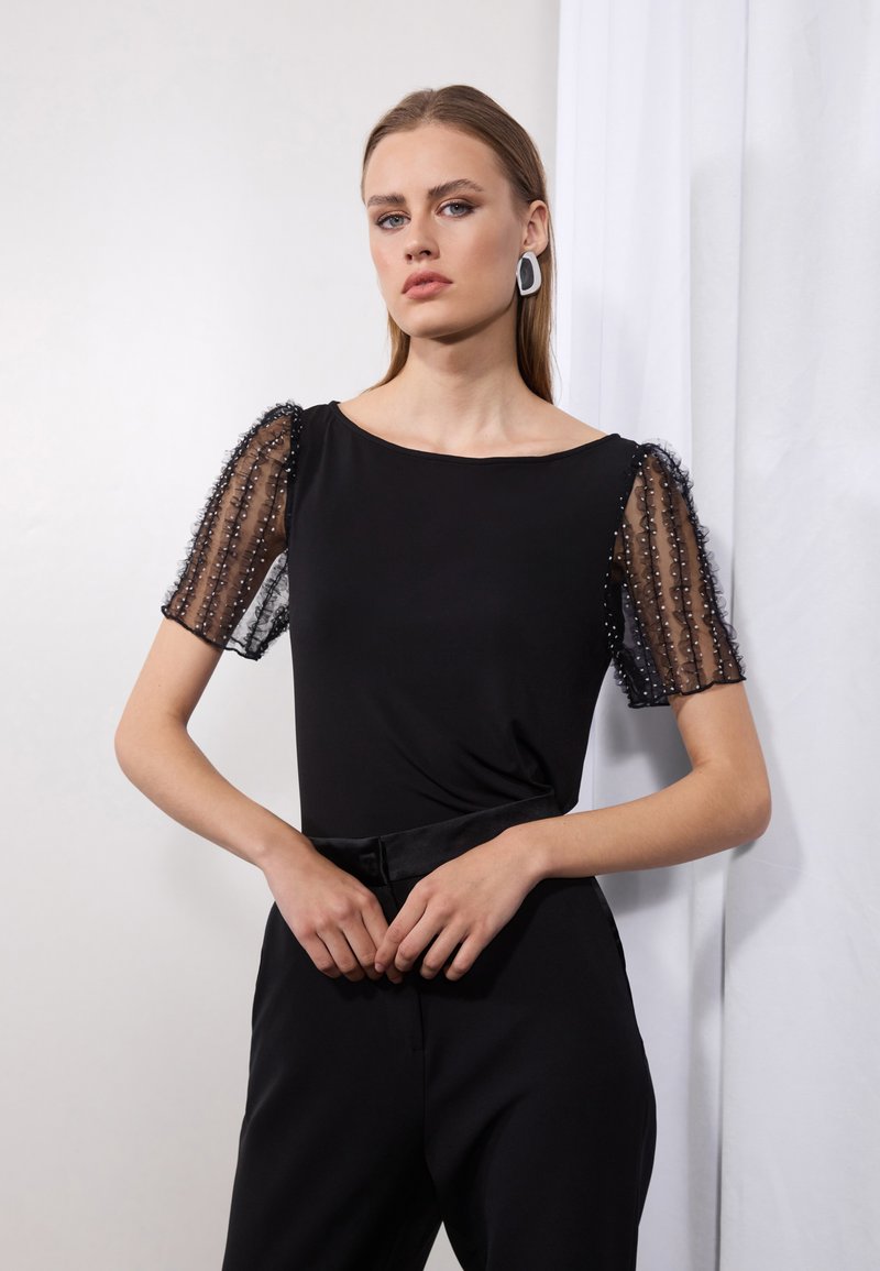 Top negru cu mâneci puff transparente decorate cu mărgele, decolteu rotund și corset ajustat. Purtați-l cu pantaloni negri cu talie înaltă.