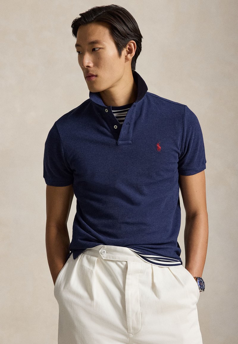 Bărbat tânăr cu păr închis la culoare poartă un tricou polo bleumarin peste un tricou cu dungi, pantaloni albi și un ceas, stând cu mâinile în buzunare și privirea spre stânga.