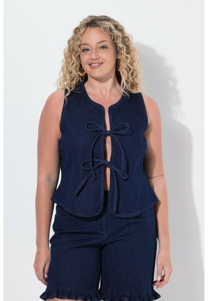 Frau mit lockigem blondem Haar, die eine ärmellose dunkelblaue Jeansweste trägt, die vorne mit zwei Schleifen gebunden ist, und passende Jeansshorts mit Rüschensaum.