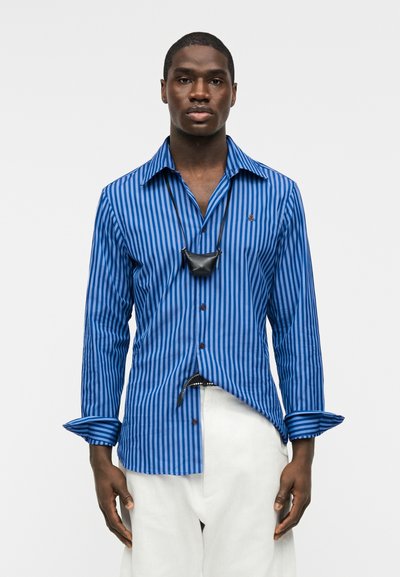 Άνδρας που φοράει μπλε πουκάμισο με κάθετες ρίγες, λευκό παντελόνι και μικρό μαύρο κρεμαστό τσαντάκι, στέκεται μπροστά σε απλό φόντο.