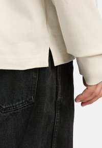 Camicia a maniche lunghe crema con fessure laterali, abbinata a pantaloni in denim neri. Il tessuto sembra morbido con una texture liscia e cuciture pulite.