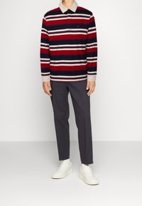Polo de manga larga a rayas en rojo, azul marino y crema, con un cuello beige y un pequeño logo. Combinado con pantalones grises oscuros y zapatillas blancas.