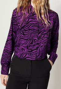 Personne portant un blouse à manches longues à motifs zèbres noir et violet, rentrée dans un pantalon noir, avec une main dans la poche.