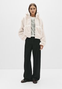 Giacca corta in pile rosa chiaro con cappuccio, tasche frontali e polsini elasticizzati; abbinata a pantaloni larghi a righe verticali neri e scarpe scure.